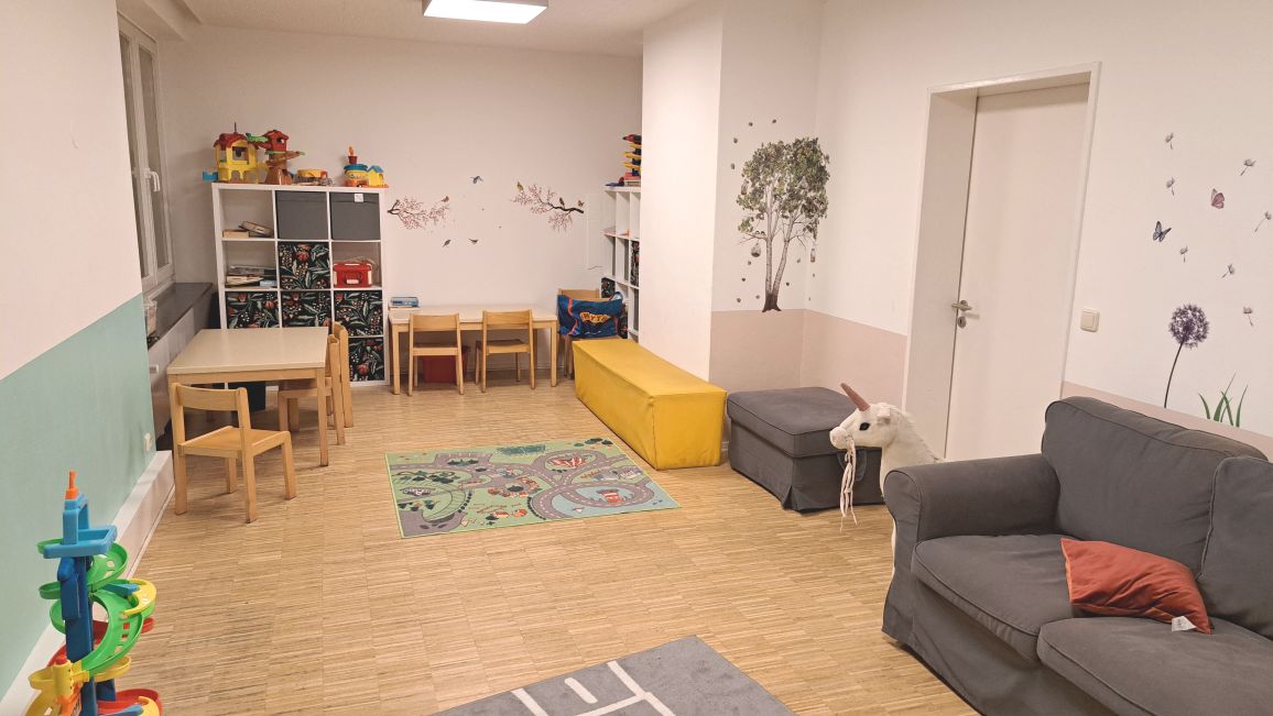 Spielzimmer mit Tischen, Stühlen, Regalen, Spielzeug und einem Sofa.